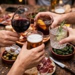 Mains en gros plan tenant des verres de vin rouge, bière et cocktail trinquent ensemble sur une table en bois rustique chargée de tapas espagnoles pour un apéro dînatoire convivial de 10 personnes.