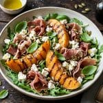 Salade estivale de melon charentais grillé, feta émiettée, tranches de jambon de Parme et graines de courge grillées, servie dans un bol en céramique sur une table en bois rustique avec de la roquette et du basilic frais.