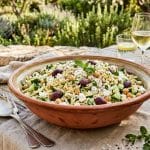 Salade d’orzo méditerranéenne avec feta, olives Kalamata, concombre et pois chiches dans un bol en terre cuite sur une table en bois en extérieur.