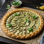 Tarte fine ronde aux courgettes disposées en rosace sur une crème de ricotta, pâte feuilletée tressée et dorée, parsemée de graines de courge et basilic sur bois sombre.